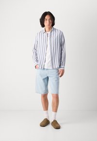 Stripet skjorte med blå og hvite striper, kombinert med lyseblå denimshorts og brune slip-on sko. Hvite sokker er synlige.