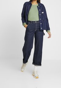 Veste à rayures bleu profond, pull vert et jean denim foncé à jambes larges, associés à des baskets claires. Superposition décontractée et contemporaine.