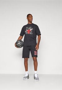 T-shirt gráfica preta com design de touro vermelho, combinada com calções pretos com a palavra "BULL" em vermelho. O modelo segura uma bola de basquete e está a usar ténis.