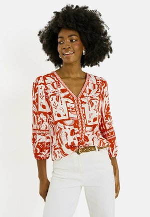 Mujer con cabello rizado que lleva una blusa con cuello en V roja y blanca estampada y pantalones blancos, sonriendo y mirando hacia un lado.