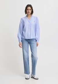 Lichtblauw knoopshirt met ruches bij de kraag en lange pofmouwen, gecombineerd met lichtblauwe denim jeans en witte sneakers.