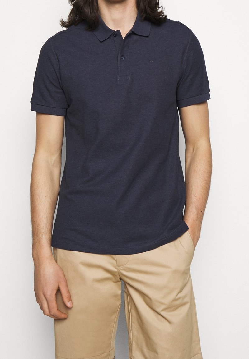 Homme portant un polo bleu marine foncé à manches courtes et un pantalon beige, debout avec une main dans la poche devant un fond uni.