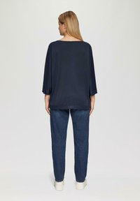 Vrouw met lang blond haar, gekleed in een losse marineblauwe top, donkere spijkerbroek en witte platform schoenen, staand met haar rug naar de camera tegen een effen achtergrond.