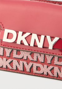 Κόκκινη τσάντα DKNY με γυαλιστερό φινίρισμα από καουτσούκ, με εμφανή ασημένια γράμματα και υφασμένο σχέδιο στο κάτω τμήμα.
