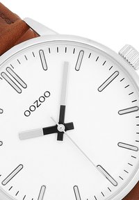 Montre avec bracelet en cuir marron, boîtier en acier inoxydable argenté, cadran blanc, aiguille des heures noire, aiguille des minutes argentée, et logo de la marque en noir.