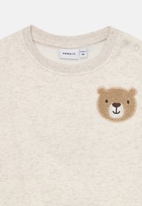 Maglietta per bambini color crema con una toppa a forma di orsetto sorridente in rilievo sul petto e bottoni a pressione su una spalla.
