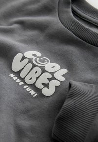 Hall hallipluus, millel on tekstuuriga trükk "COOL VIBES" ja naeratav nägu. Soonikkoes mansetid lisavad disainile detaili.