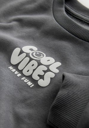 Šedá mikina s texturovaným potiskem s nápisem "COOL VIBES" a usměvavým obličejem. Žebrované manžety dodávají celkovému designu detail.