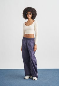 Γυναίκα με σγουρά μαλλιά που φοράει γυαλιά ηλίου, μπεζ crop top με μακριά μανίκια, χαλαρό μωβ παντελόνι και λευκά αθλητικά παπούτσια, στέκεται σε μπλε χαλί.