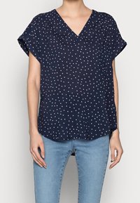 Blouse bleu marine à manches courtes avec un col en V, ornée de pois blancs, à coupe décontractée et ourlet arrondi. Fabriquée en tissu léger.