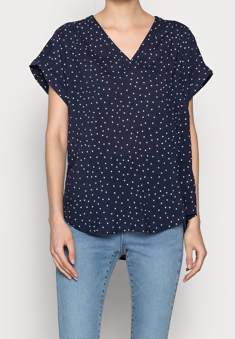 Blouse bleu marine à manches courtes avec un col en V, ornée de pois blancs, à coupe décontractée et ourlet arrondi. Fabriquée en tissu léger.