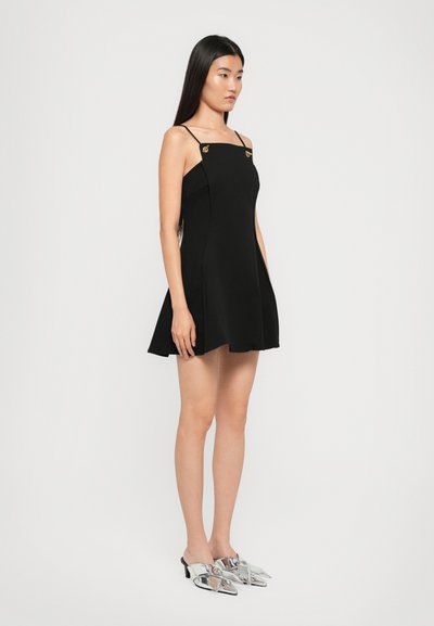 Robe noire sans manches avec une silhouette évasée, présentant des accents dorés sur les bretelles. Le mannequin porte des mules argentées à talons, posant devant un fond blanc.