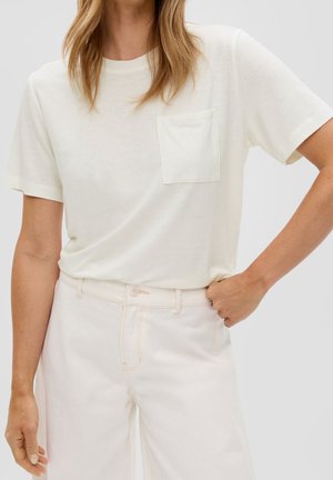 Femme portant un t-shirt blanc uni à manches courtes avec une poche poitrine et un pantalon taille haute rose pâle, main dans la poche.