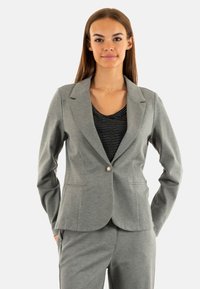 Blazer gris de tela suave con solapas de muesca, cierre de un solo botón y ajuste estructurado. Combinado con una blusa de rayas en blanco y negro.