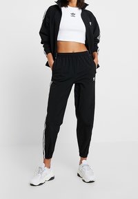 Svart träningsoverall med vita tre-stripes accenter, bestående av en zip-upp jacka och smala byxor. Kombinera med chunky vita sneakers.