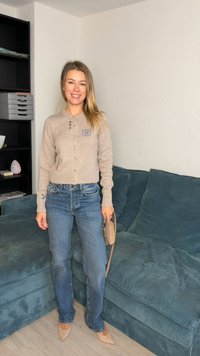 Beige ribgebreid vest met knoopsluiting en bloemenborduursel, gecombineerd met lichtblauwe spijkerbroek. Tan puntige platte schoenen en een beige handtas.