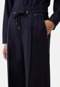 Donkerblauwe broek met elastische tailleband, trekkoord, zijzakken en een hand gedeeltelijk in de rechterzak, gecombineerd met een bijpassend bovenstuk.