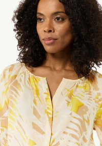 Leichte, gemusterte Bluse in Gelb- und Beigetönen, mit einem runden Ausschnitt und sanften Falten nahe dem Kragen.