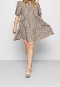 Kobieta spacerująca w beżowej sukience w kwiaty z krótkim rękawem i beżowych sandałach na odkrytej pięcie na jednolitym jasnym tle.