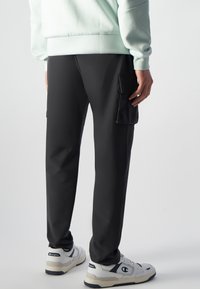 Zwarte cargobroek met zijzakken, gladde textuur en een tapered fit, gecombineerd met witte sneakers en een lichtblauwe sweatshirt.