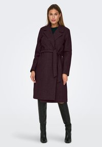 Femme portant un manteau en laine violet foncé ceinturé jusqu'aux genoux et des bottes noires jusqu'aux genoux, se tenant devant un fond gris clair uni.