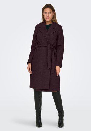 Femme portant un manteau en laine violet foncé ceinturé jusqu'aux genoux et des bottes noires jusqu'aux genoux, se tenant devant un fond gris clair uni.