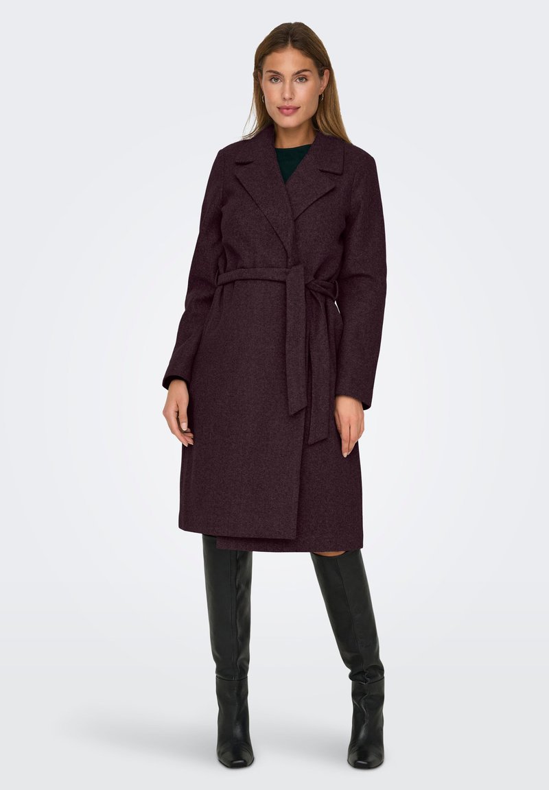Femme portant un manteau en laine violet foncé ceinturé jusqu'aux genoux et des bottes noires jusqu'aux genoux, se tenant devant un fond gris clair uni.