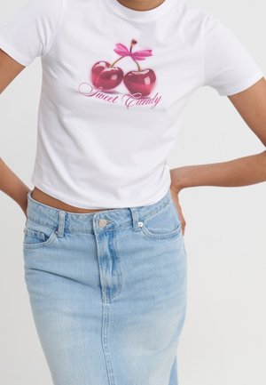 Kobieta ubrana w biały krótki t-shirt z czerwonymi wiśniami i tekstem "Sweet Candy", zestawiony z jasnoniebieską, wysoką spódnicą jeansową.