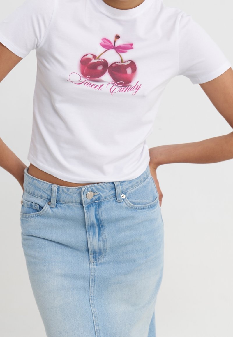 Femme portant un t-shirt court blanc avec des cerises rouges et le texte "Sweet Candy", associé à une jupe en jean taille haute bleu clair.