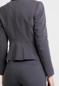 Blazer gris sur mesure avec un design péplum, texture lisse, épaules structurées et motif discret à fines rayures, doté d'une couture arrière et sans quincaillerie visible.