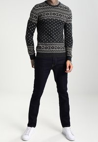Maglione scuro con motivo a fantasie bianche geometriche, abbinato a jeans denim scuri e sneakers bianche. Il maglione presenta un colletto e un orlo a coste.