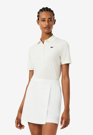 Femme aux cheveux tressés portant une chemise blanche à manches courtes avec col et une jupe portefeuille blanche sur un fond uni.