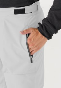 Lichtgrijze cargobroek heeft een ritszak met een zwarte ritslip, een klittenbandsluiting in de taille en een gladde, gestructureerde stof.
