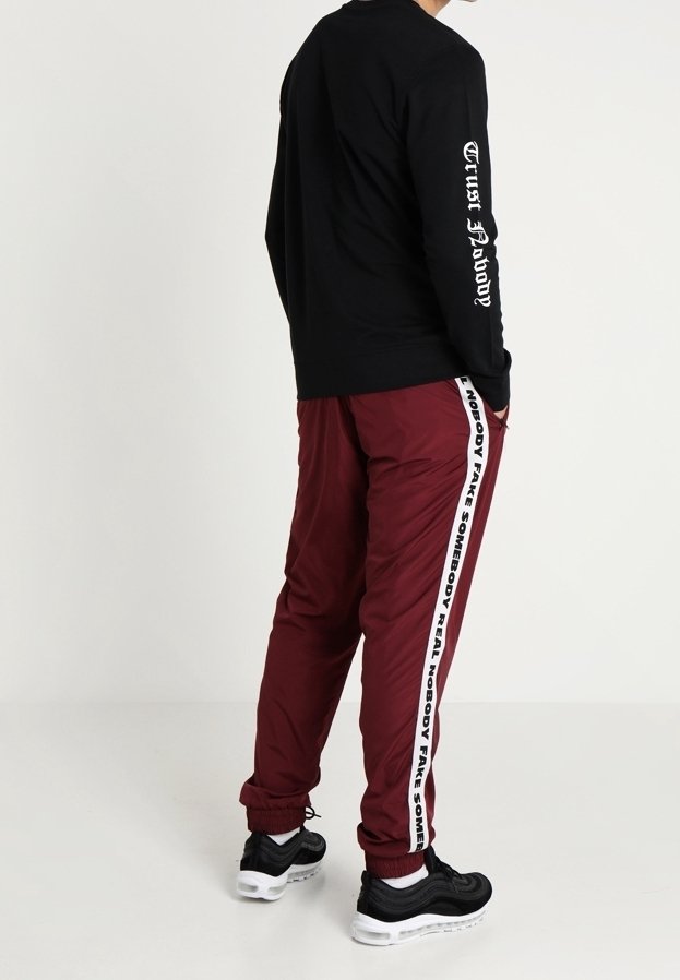 Homme debout les mains dans les poches, portant un sweat-shirt noir avec l'inscription "Trust Nobody" sur la manche et un pantalon bordeaux avec une bande latérale blanche et un texte.