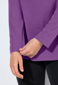 Fialová dlouhá rukávová košile vyrobená z měkké látky, s kulatým výstřihem a mírně přiléhavým manžetou; viditelné ruce, jak upravují rukáv.