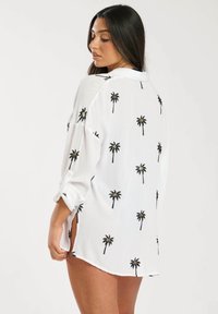 Camisa blanca de botones con estampado de palmeras negras, con mangas enrollables y aberturas laterales para mayor movimiento y estilo. Tejido ligero.
