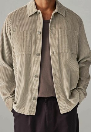 Homme portant une chemise beige en velours côtelé boutonnée par-dessus un t-shirt marron foncé, les mains dans les poches d'un pantalon sombre, sur un fond neutre.