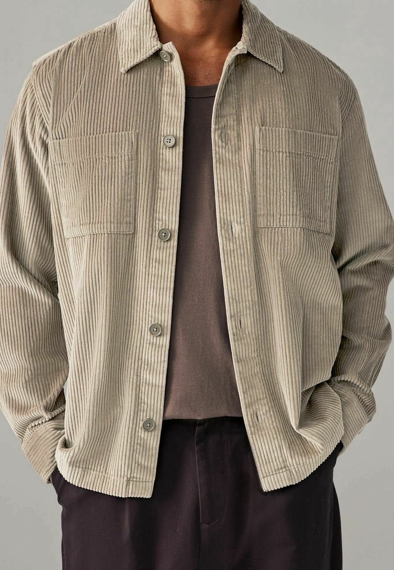 Homme portant une chemise beige en velours côtelé boutonnée par-dessus un t-shirt marron foncé, les mains dans les poches d'un pantalon sombre, sur un fond neutre.