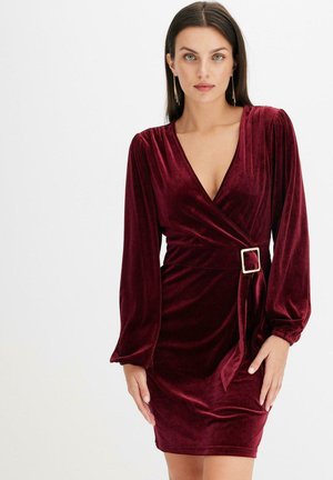 Vestito avvolgente in velluto burgundy con maniche lunghe, scollatura a V e cintura in tessuto con dettaglio della fibbia quadrata. Texture liscia in tutto il vestito.