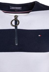 Granatowo-biały sweter w paski z zamkiem błyskawicznym, wyposażony w okrągły metalowy suwak, naszywkę z logo oraz kontrastowe faktury.