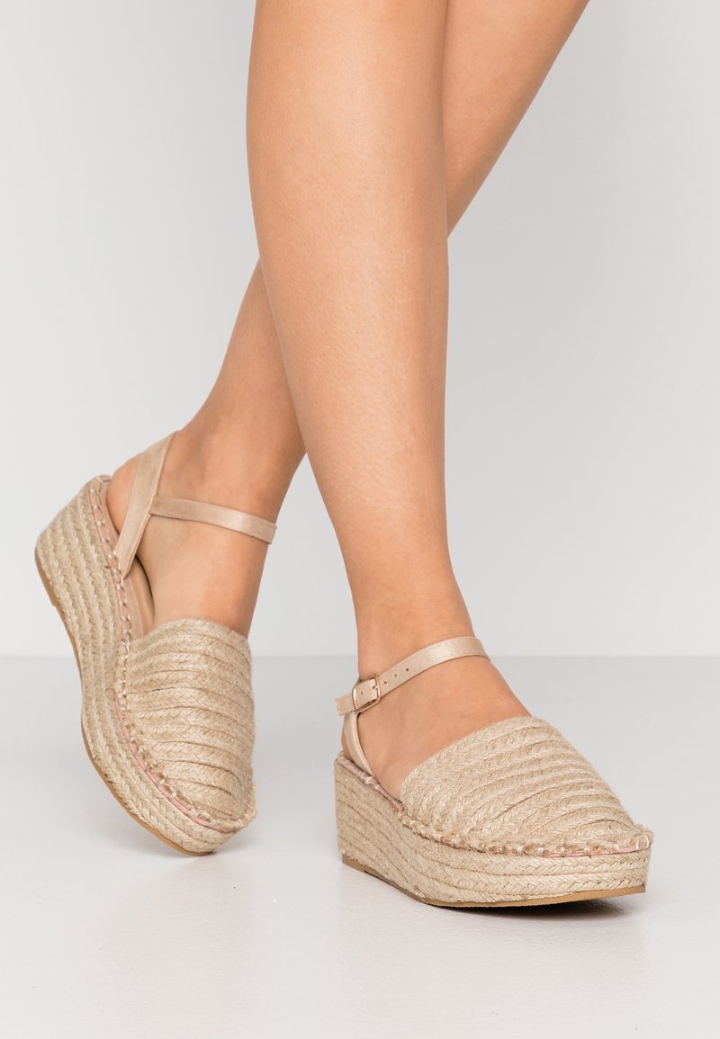 Jambes portant des sandales compensées en espadrille beige avec des brides chevilles, motif tressé texturé, debout sur un sol et un fond gris clair.