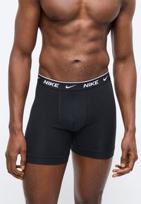 Μυώδης άνδρας που φοράει μαύρα εσώρουχα Nike boxer με το λογότυπο στη μέση, στέκεται με τα χέρια χαλαρά μπροστά σε απλό φωτεινό φόντο.