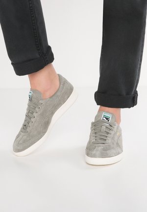 Person iført grå ruskind Puma sneakers med hvide såler og grønt logo, kombineret med op rullede sorte jeans mod en ensfarvet baggrund.