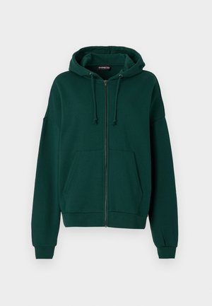 Haut à capuche zippé vert foncé en tissu doux. Il dispose d'une capuche avec cordon de serrage, de poches avant et de poignets et un bas côtelés.