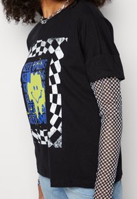 T-shirt nera a maniche corte con una grafica di una faccina sorridente gialla con testo blu e un accentuato motivo a scacchi. Sotto, indossata una camicia a maniche lunghe a scacchi.