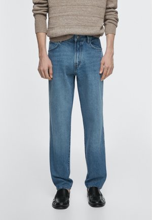 Person trägt lockere blaue Jeans, schwarze Schuhe und einen beigen Strickpullover, steht vor einem schlichten, hellen Hintergrund.