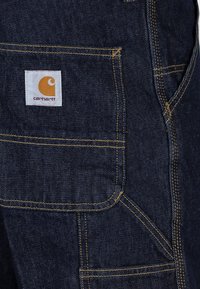 Overoles de mezclilla azul con costuras amarillas y un parche del logotipo de Carhartt visible en el bolsillo delantero. Tela duradera con costuras precisas.