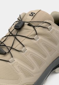Beige tursko for hiking med hurtigløselige svarte elastiske snører og en polstret mesh-tunge med en svart logo.