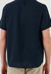 Granatowa koszula z krótkim rękawem wykonana z lekkiego materiału, z okrągłym dekoltem i prostym krojem z lekko zaokrąglonym dołem.