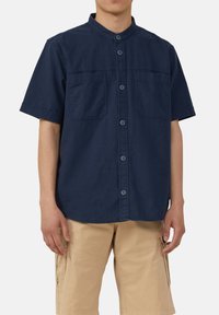 Marineblauw overhemd met korte mouwen en staande kraag, voor sluiting met knopen, twee borstzakken en een zachte katoentextuur. Gecombineerd met beige cargoshorts.